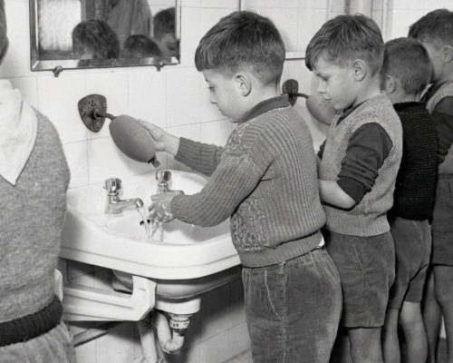 Des jeunes garçons (écoliers) se lavant les mains à un lavabo commun à l'aide du savon d'écolier fixé au mur